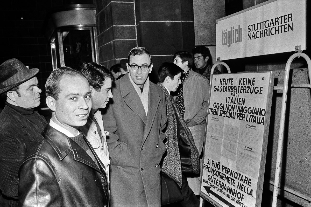 Zu sehen sind Männer, die vor einem Schild stehen, welches darauf hinwiest, dass an diesem Tag keine Gastarbeiter Züge nach Italien fahren.