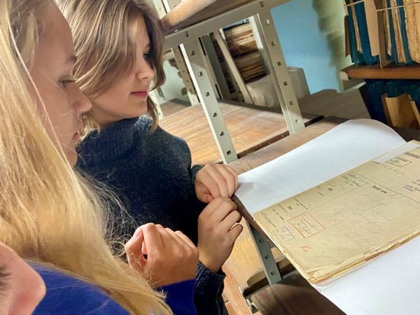 Zwei Mädchen (links im Bild) stehen im Archivmagazin vor einem Regal (rechts im Bild) und sehen eine darauf liegende Akte an.