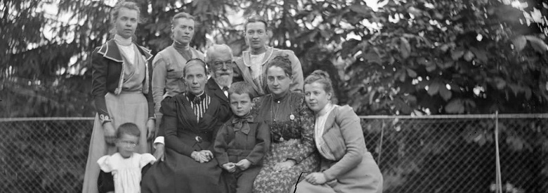 Zu sehen ist ein schwarz weiß Bild. Acht Erwachsene und zwei Kinder sitzen eung beisammen, der Kamera zugewandt und posieren für das Bild. Sie befinden sich auf einer Terasse oder Balkon, denn hinter dem Zaun, vor dem die Familie steht, sind Baumkronen.