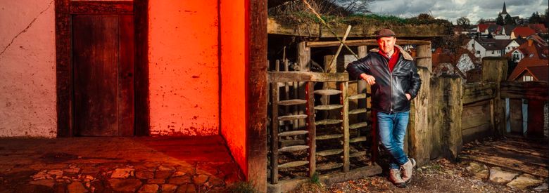 Ausschnitt aus Farbfoto von Karl Banghard, der auf der rechten Seite des Bilds angelehnt gegen einen alt aussehenden Holzzaun steht. Auf der linken Seite ein rot angestrahltes, sehr alt wirkendes Haus.