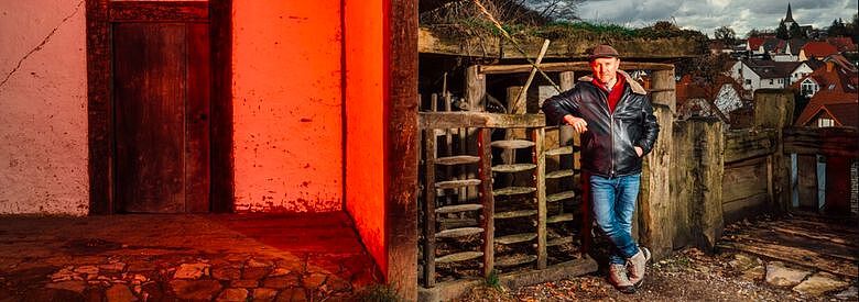 Ausschnitt aus Farbfoto von Karl Banghard, der auf der rechten Seite des Bilds angelehnt gegen einen alt aussehenden Holzzaun steht. Auf der linken Seite ein rot angestrahltes, sehr alt wirkendes Haus.