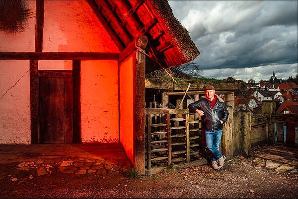 Farbfoto von Karl Banghard, der auf der rechten Seite des Bilds angelehnt gegen einen alt aussehenden Holzzaun steht. Auf der linken Seite ein rot angestrahltes, sehr alt wirkendes Haus.