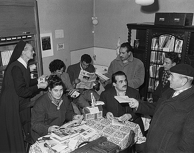 Zu sehen sind mehrere Menschen in einem kleinen Raum um einen Tisch herum verteilt. Sie hantieren mit Briefen und Zeitungen.