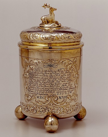Zu sehen ist ein goldener Becher mit drei runden Füßen. Auf dem Deckel thront ein liegender Hirsch. Auf dem Becher selber ist ein Text eingraviert, der von Ranken verziert wird.