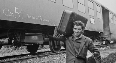 Zu sehen ist ein Mann, der einen Koffer trägt. Hinter ihm ist ein Zug.
